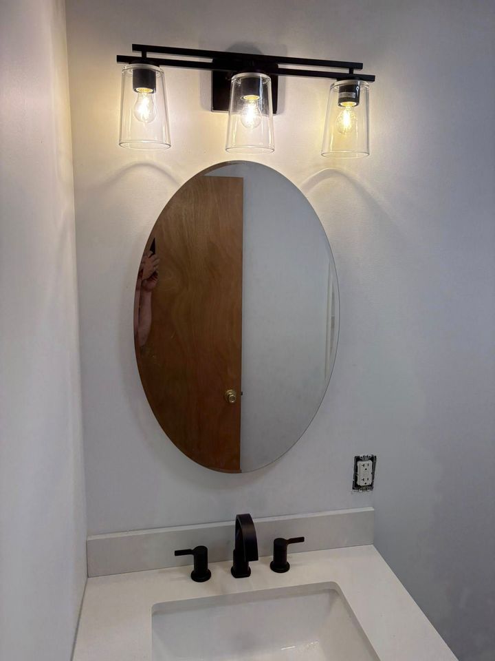 Bathroom with black fixtures, oval mirror, and three-light sconce over a white countertop with a sink.