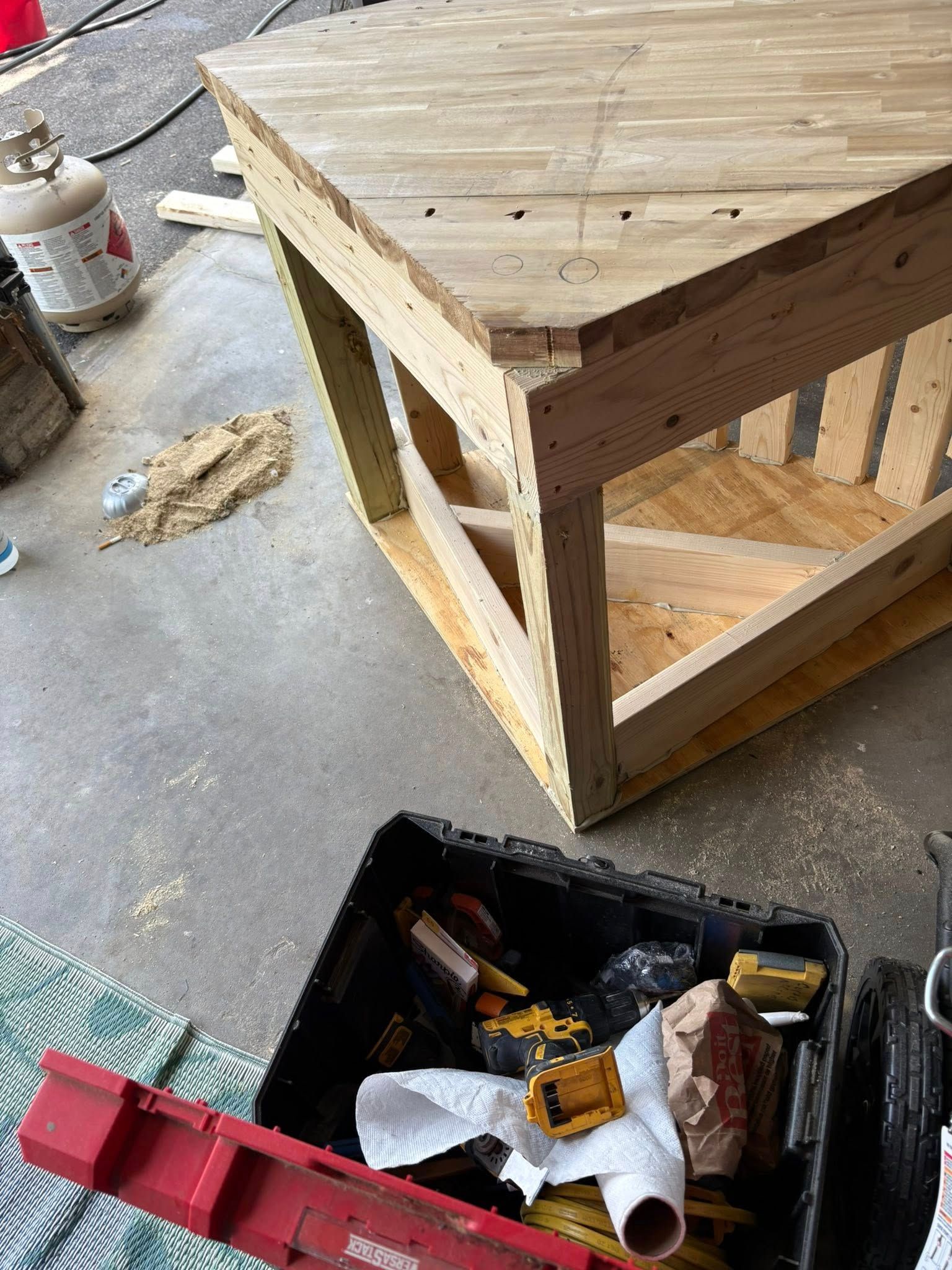 Wooden table under construction, tools in a black bin, propane tank in background, concrete floor.