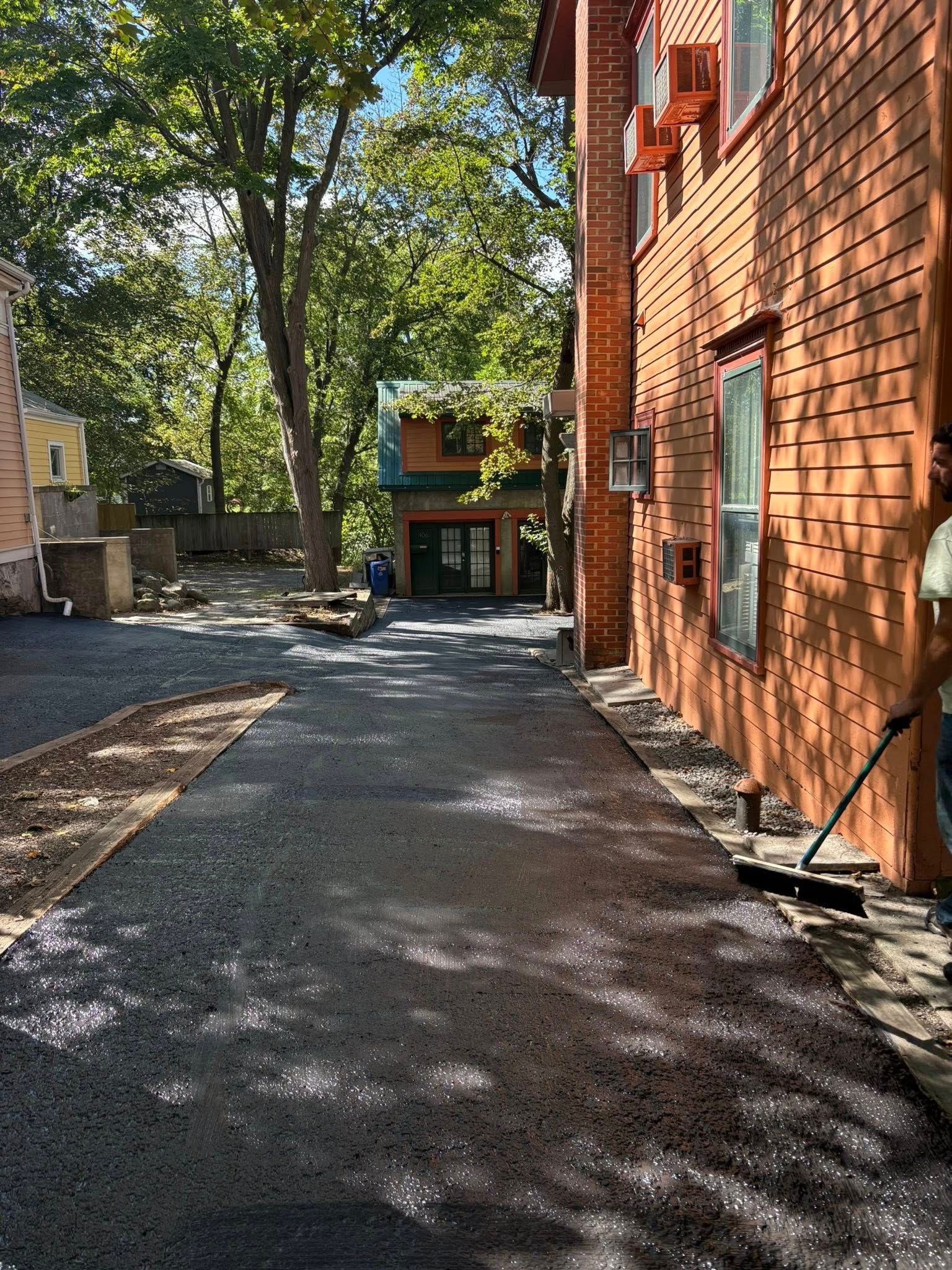 Fresh asphalt driveway next to a brick building; worker sweeping.