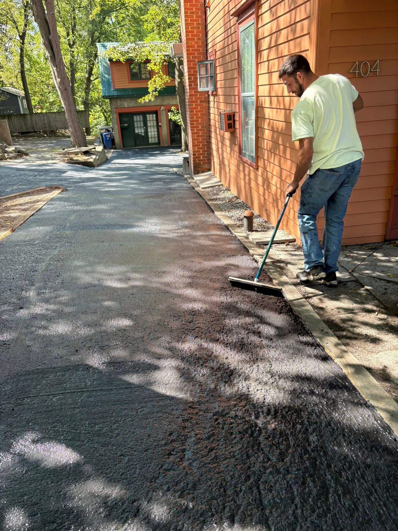 Man using a squeegee to smooth fresh asphalt on a driveway next to a building.