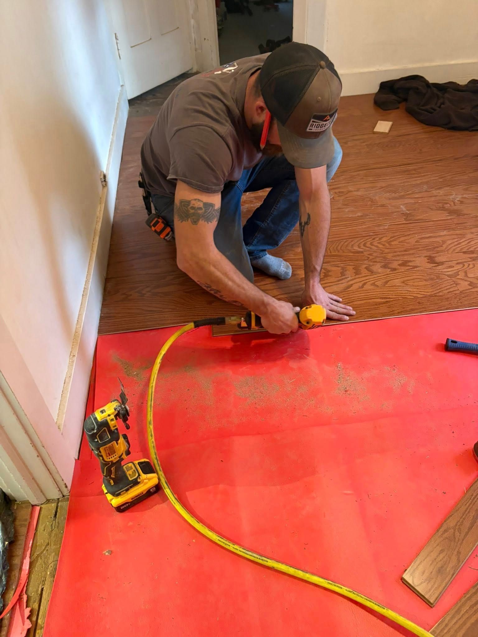Person installing flooring, kneeling with nail gun. Red underlayment visible, yellow air hose connected.