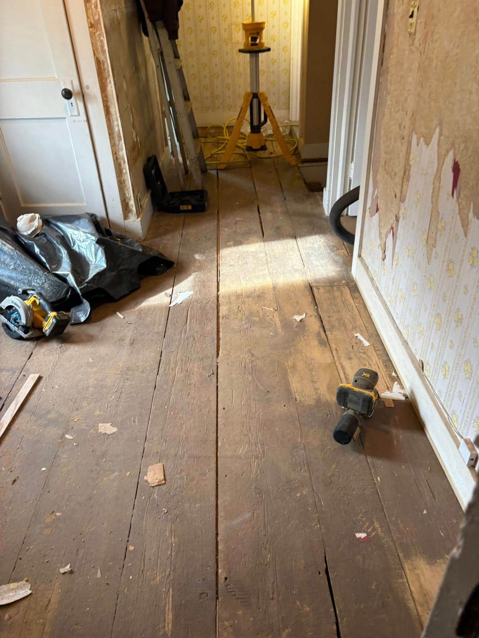 A hallway under renovation, with exposed wooden floorboards, tools, and a laser level on a tripod.