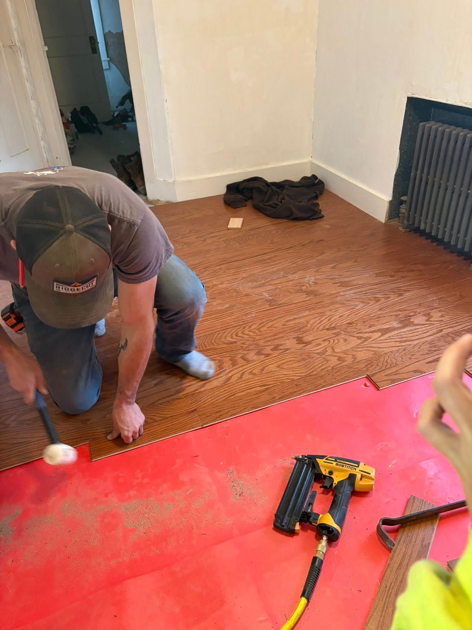 Person kneeling on floor installing flooring with hammer, air nailer, in room with white walls.