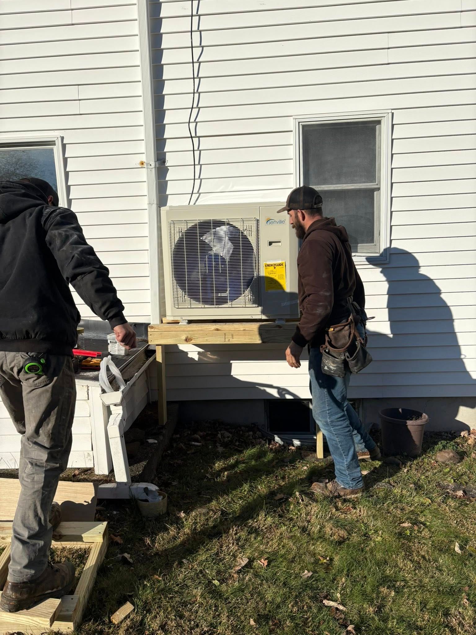 Two workers installing an air conditioning unit on a wooden platform against a white house.