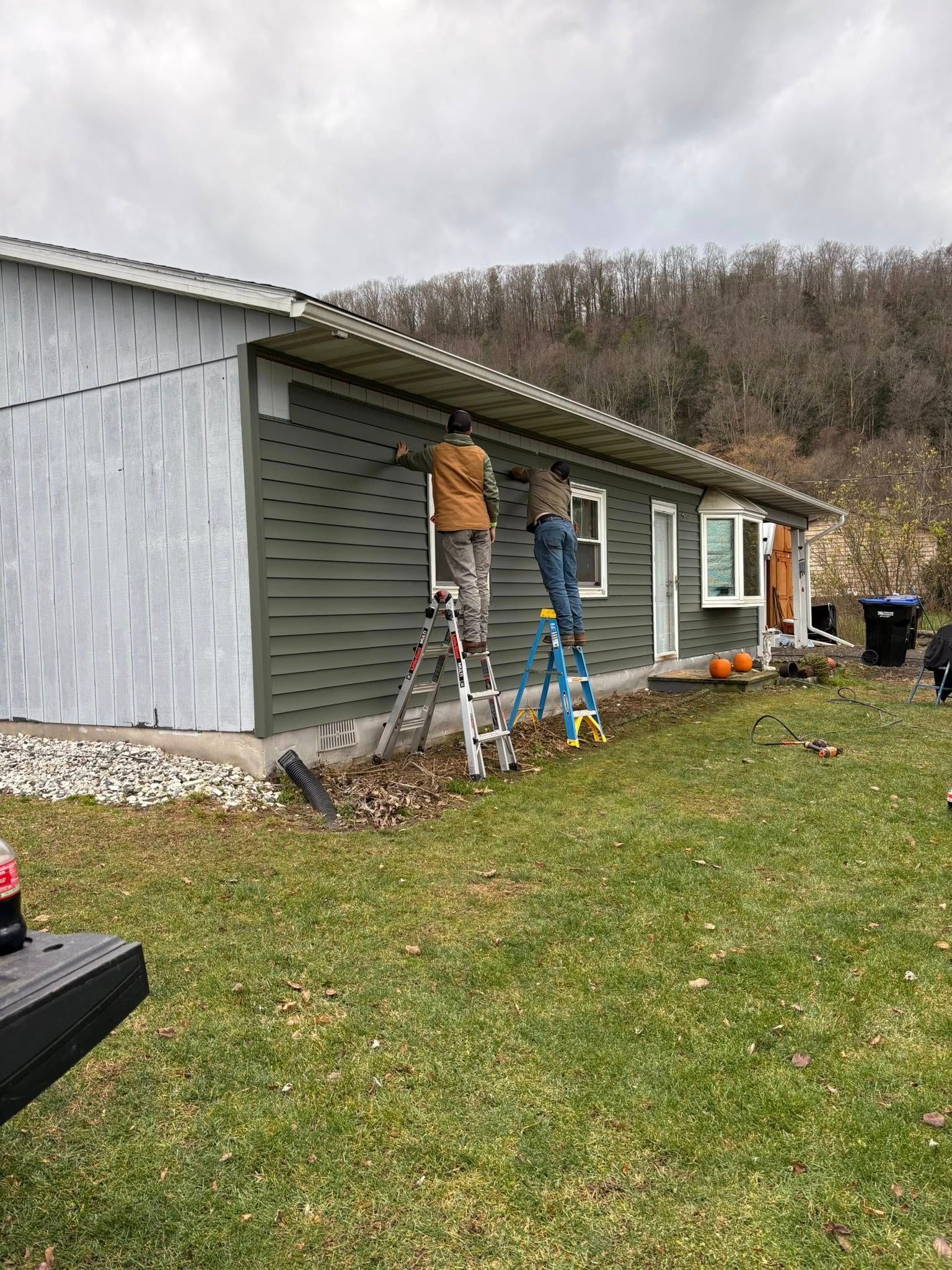 Two people installing green siding on a house. One is on a gray ladder, the other on a blue one. Overcast day.