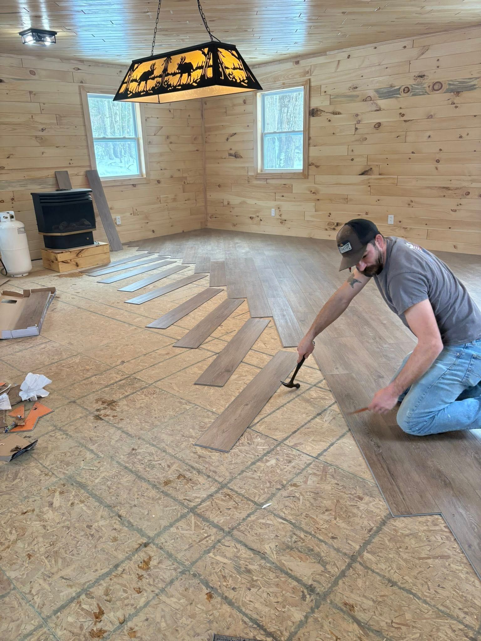 Man kneeling, installing wood flooring in a room with a fireplace and windows; interior.