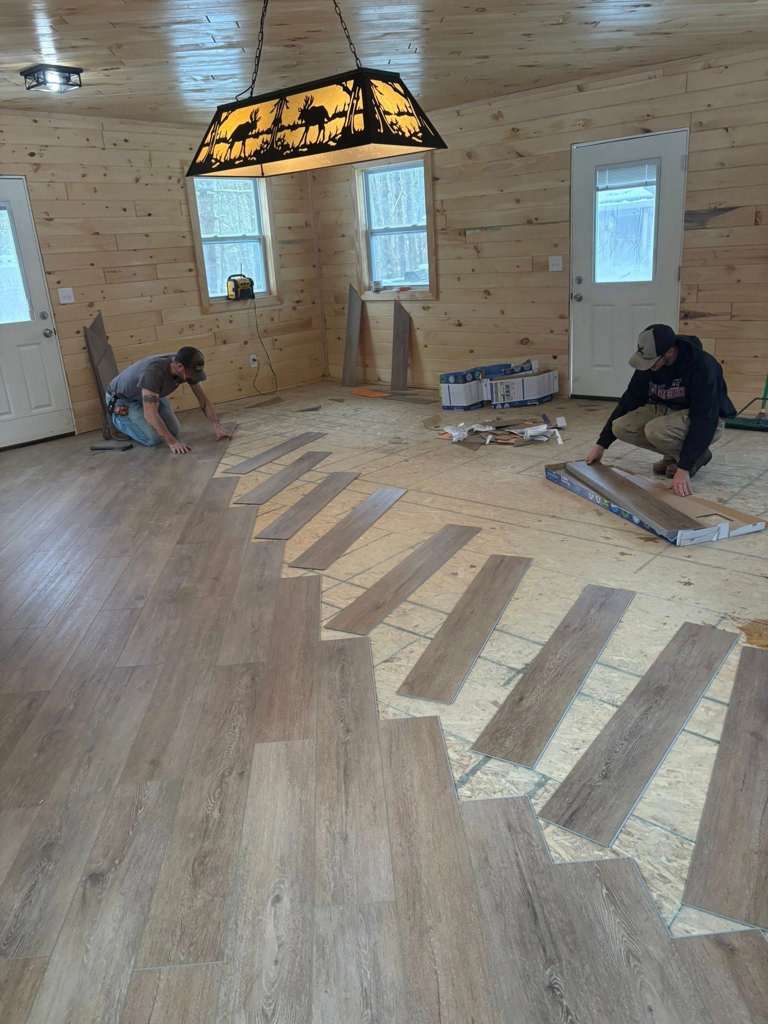 Two workers installing wood-look flooring in a room with wood-paneled walls and a hanging light fixture.