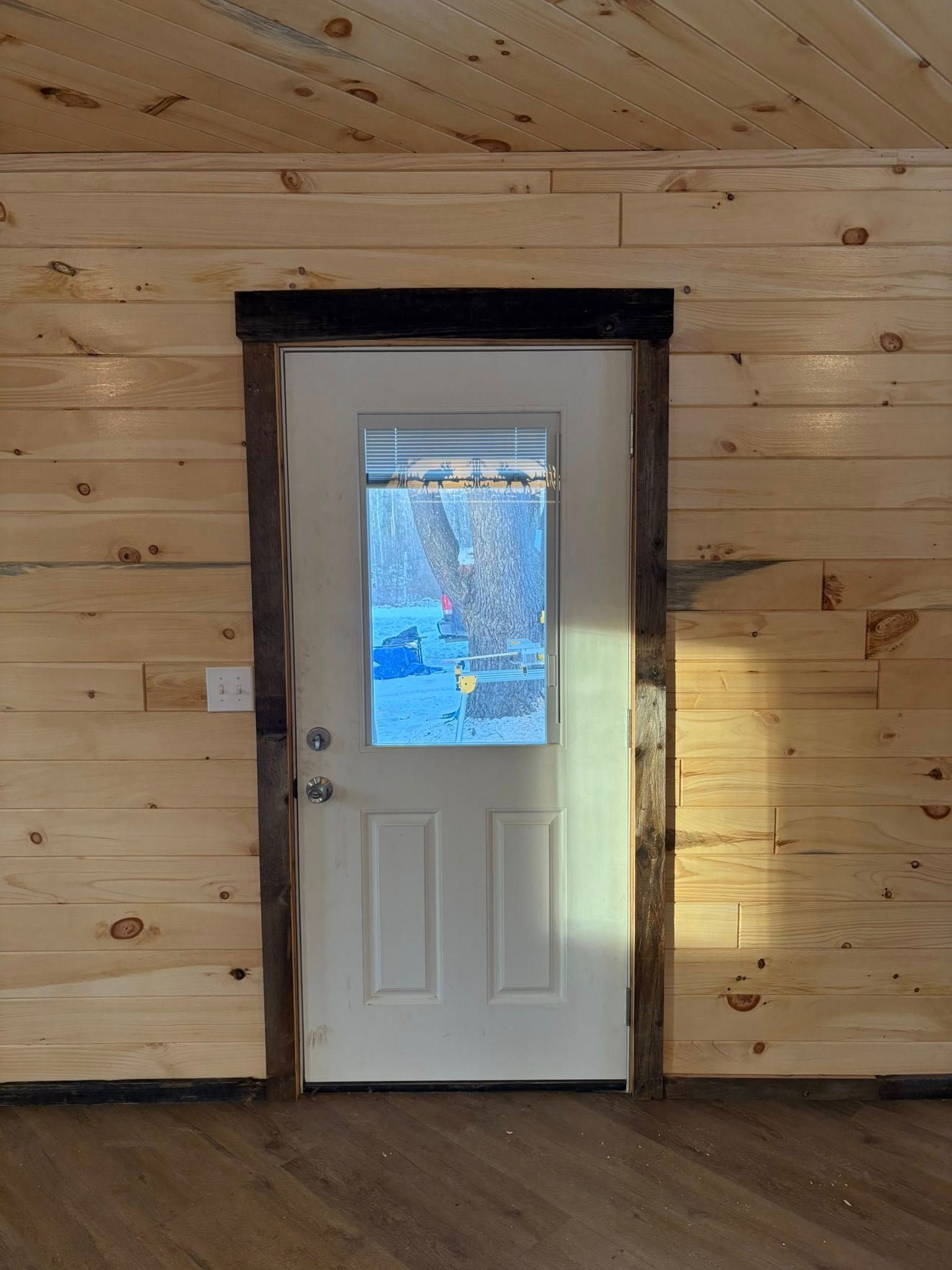 White door with window, surrounded by dark wood trim, set in a wooden paneled wall.