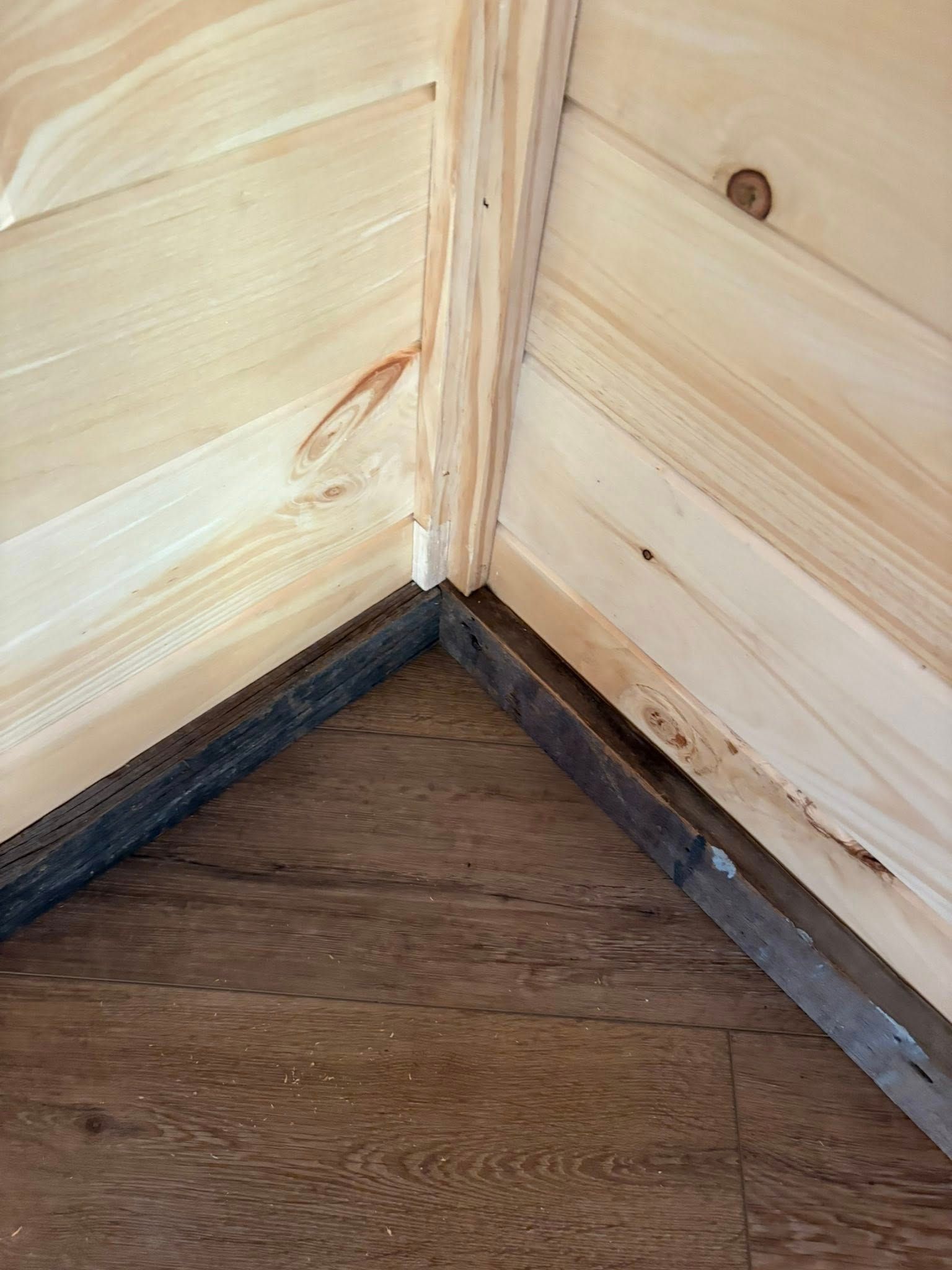 Corner of a wooden structure: walls and floor meet at a sharp angle. Light wood walls, dark floor.