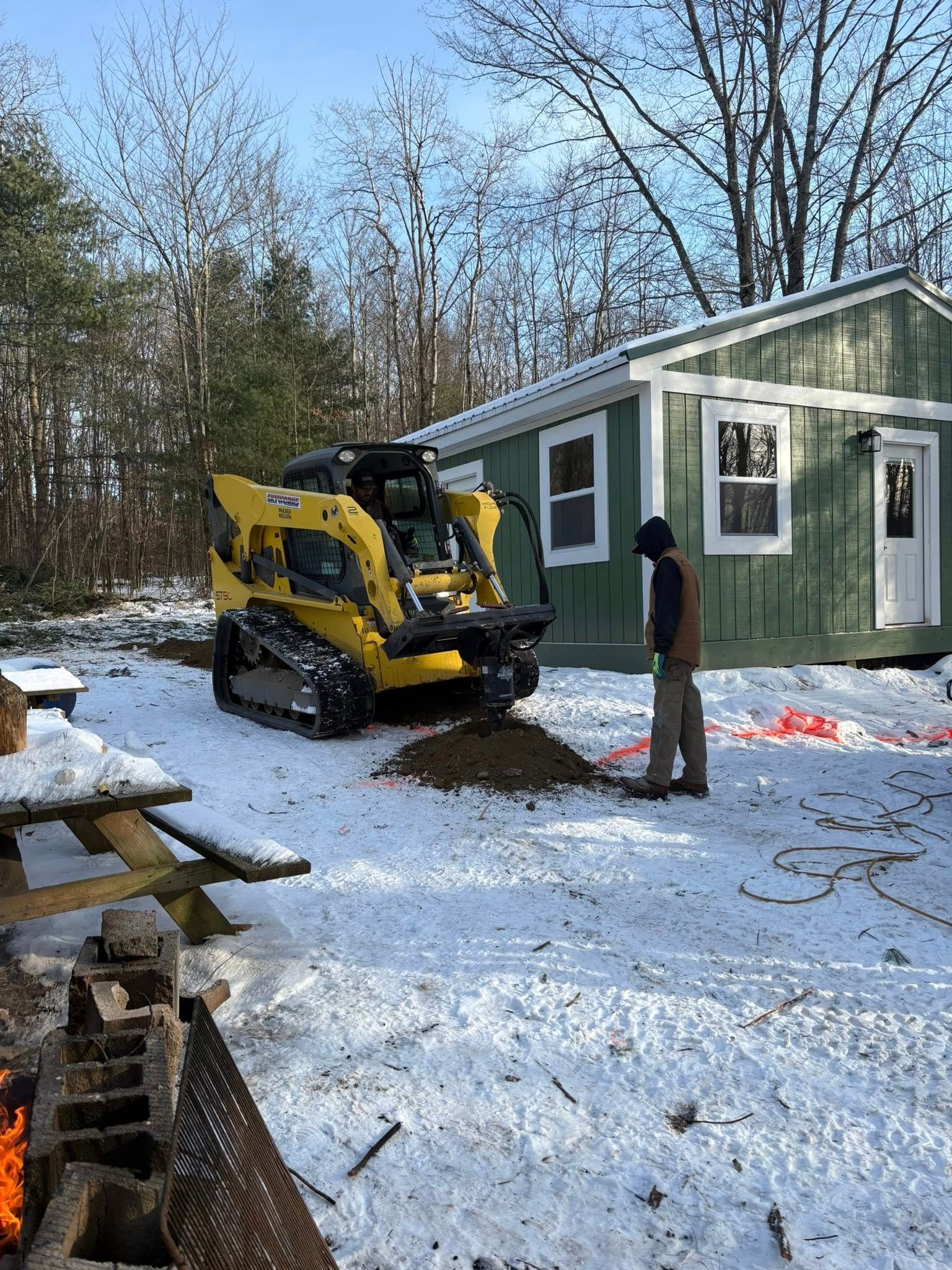 Yellow skid steer and worker near a green cabin in a snowy yard.