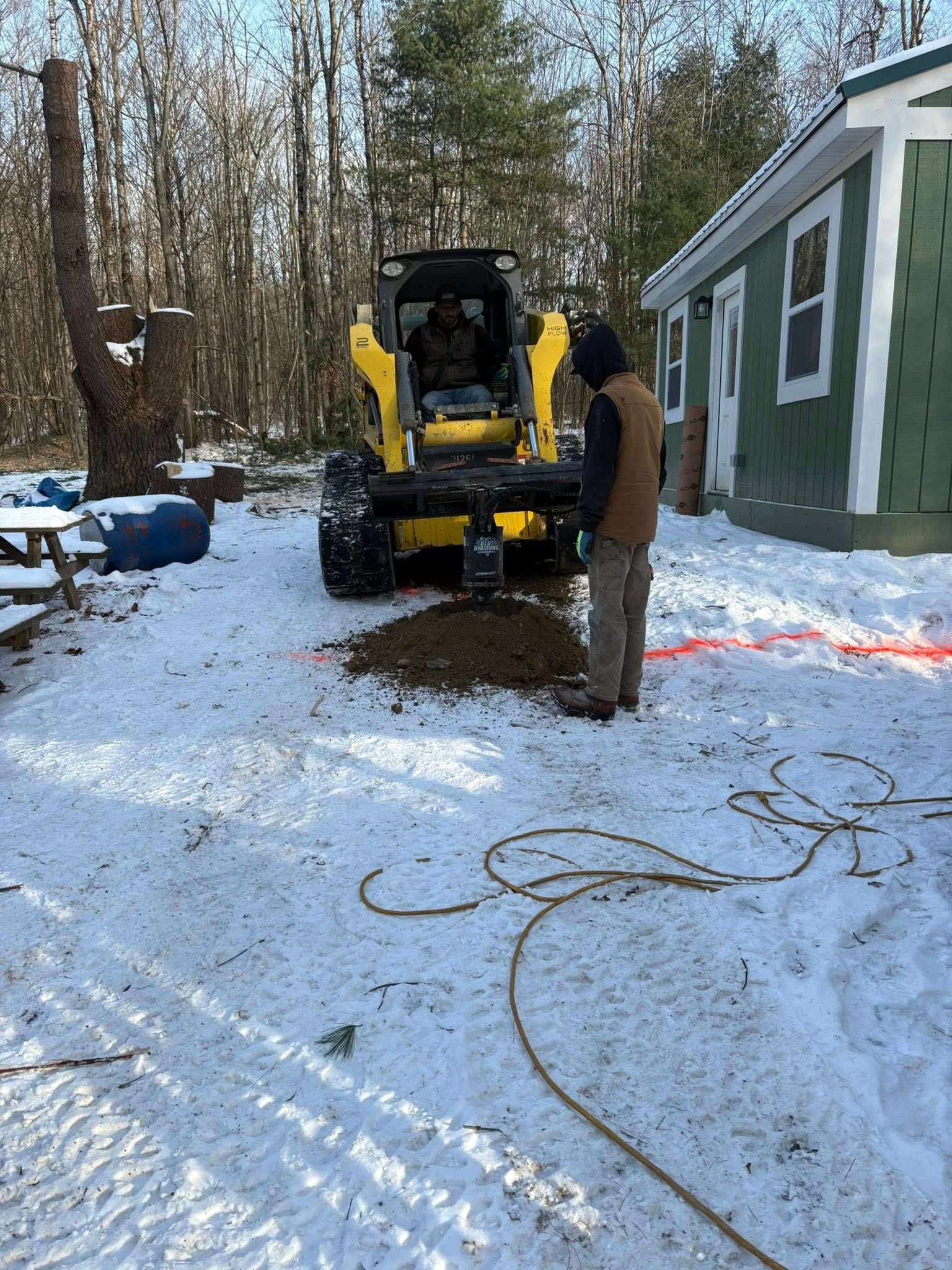 Person watches a yellow skid steer drill into snowy ground near a building.