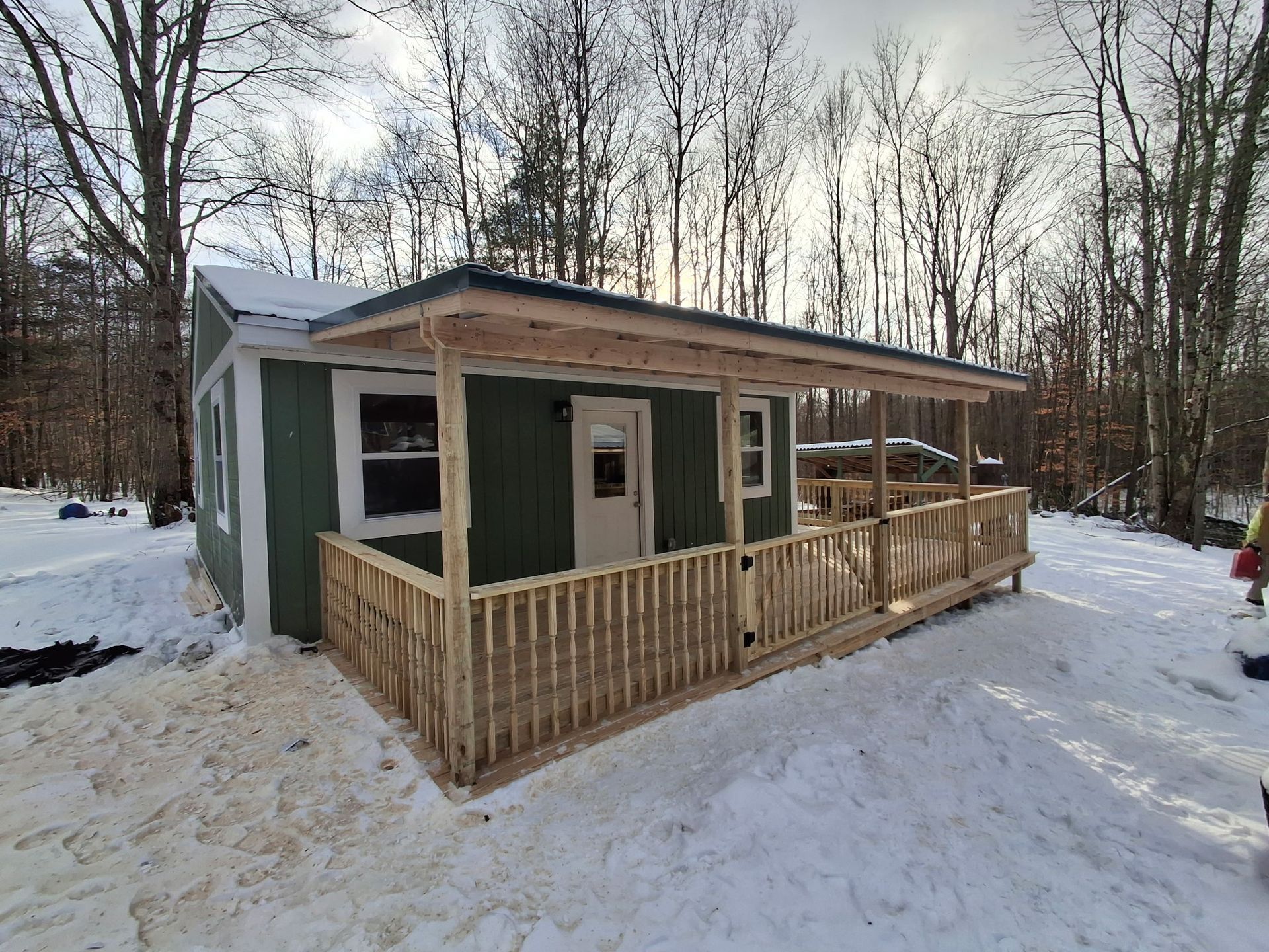 Green cabin with porch and snow in wooded area.