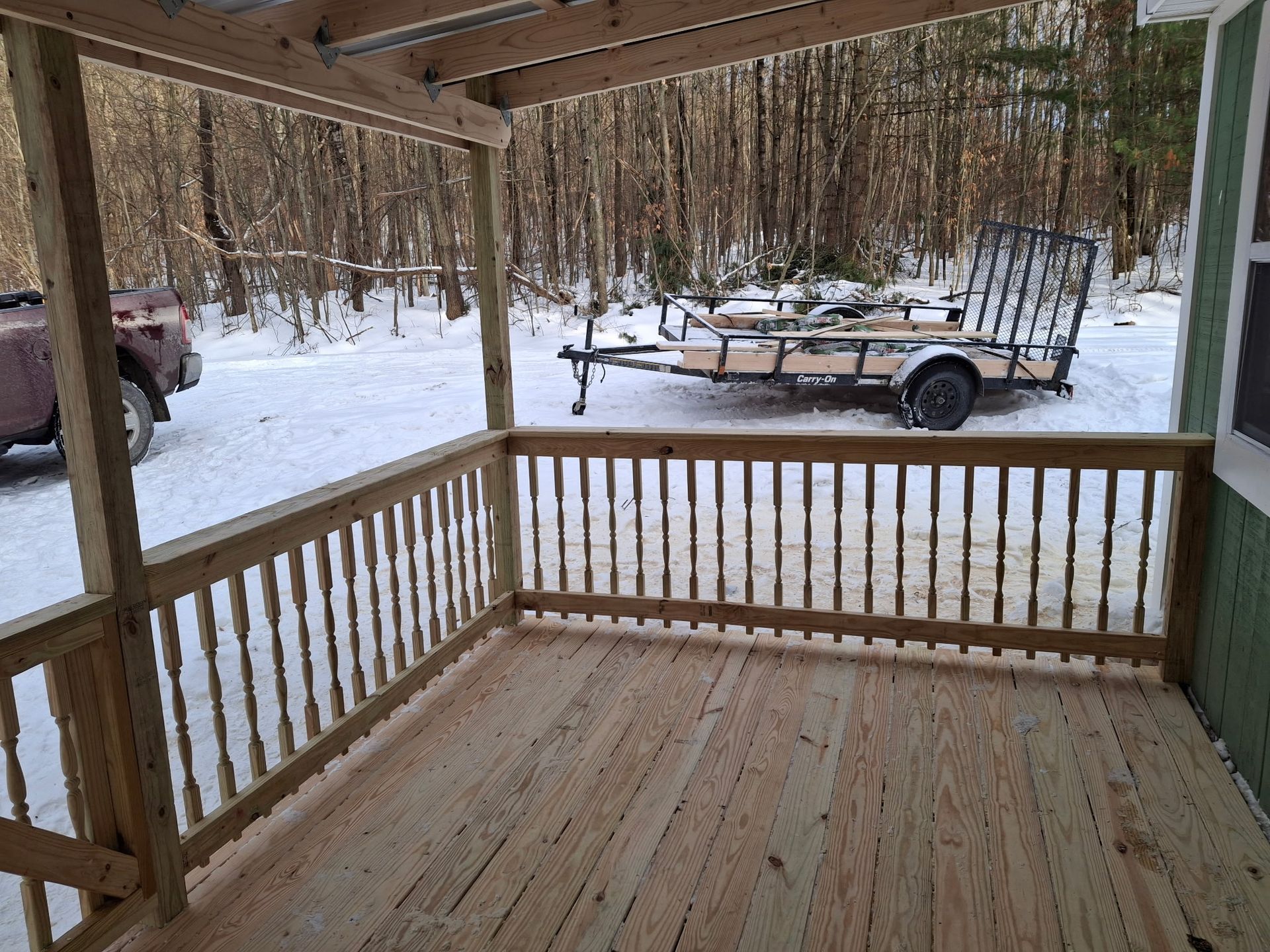Wooden porch with railing, looking out onto a snowy landscape with a trailer and truck.