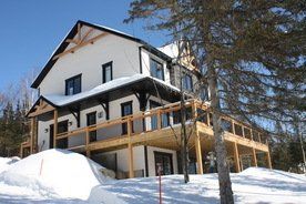 Une grande maison blanche avec une terrasse en bois est entourée de neige et d'arbres.