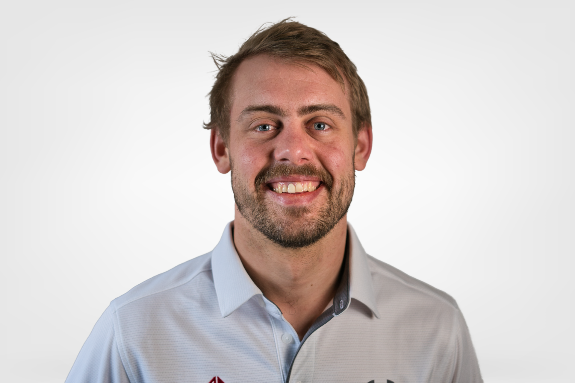 Man with a beard smiling, wearing a light blue collared shirt, against a white background.