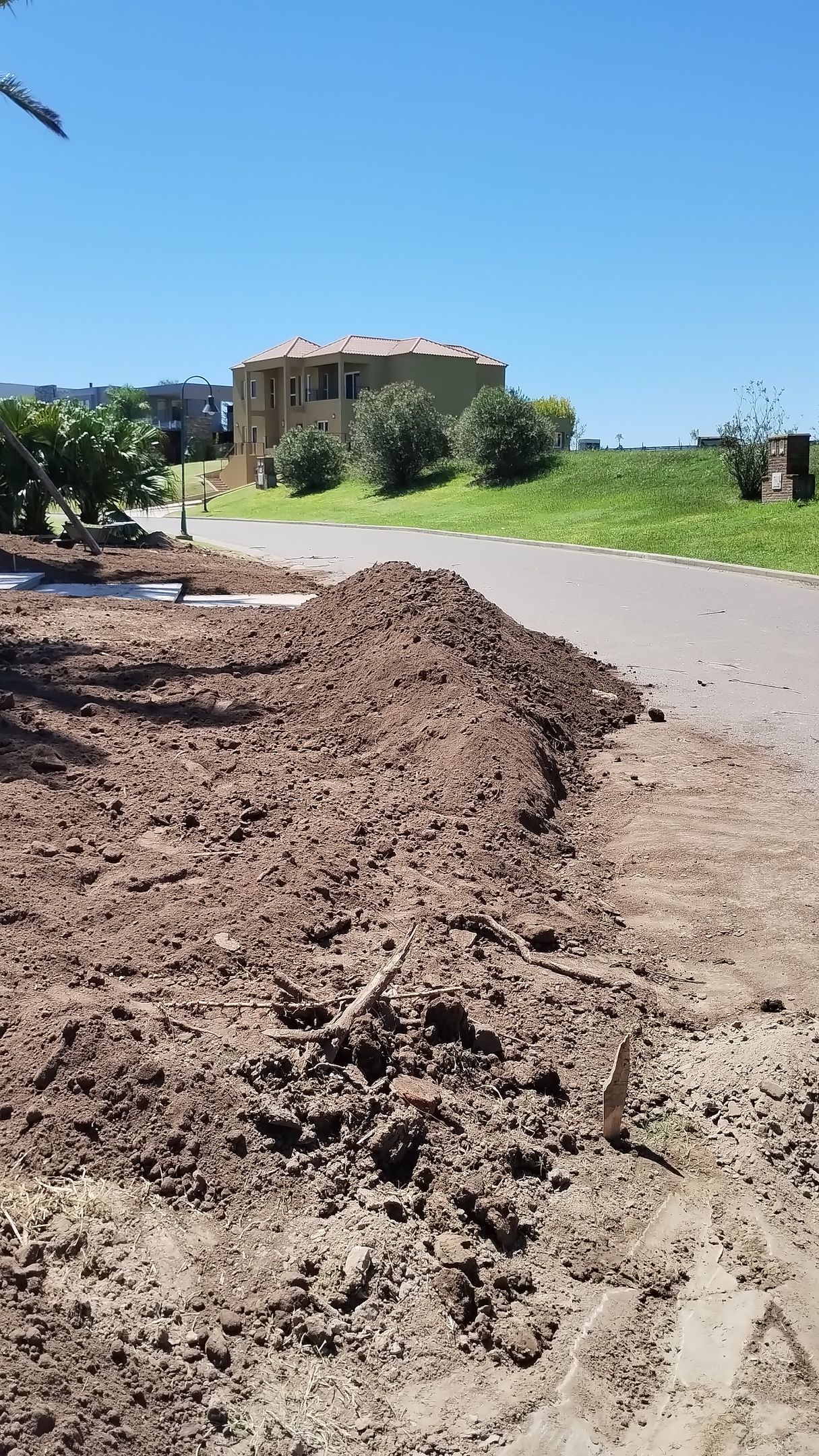 Pila de mantillo oscuro en un camino de tierra, con un edificio y árboles al fondo bajo un cielo azul.