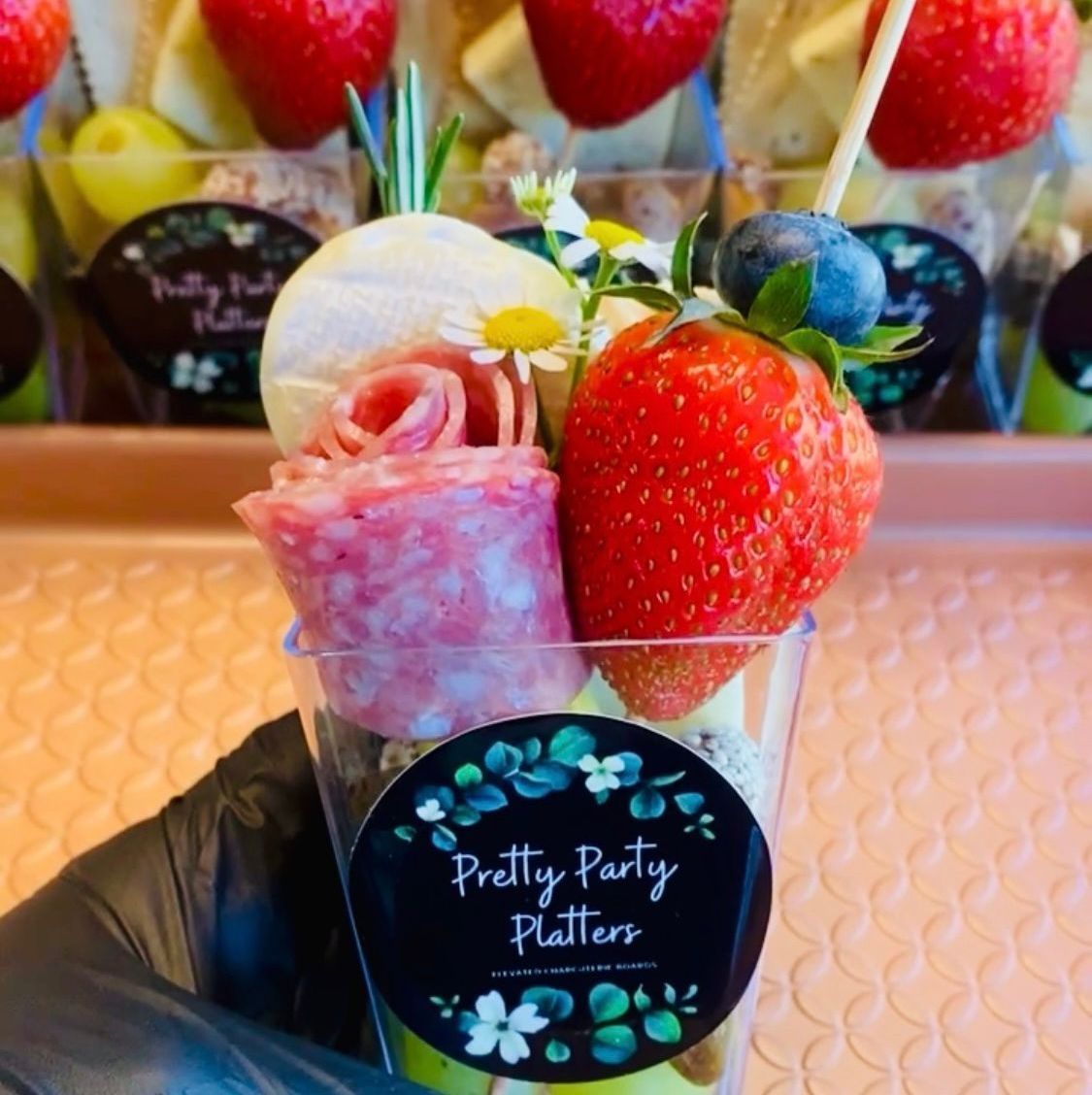 Hand holding a clear cup with salami rose, strawberry, blueberry, and garnish from Pretty Party Platters.