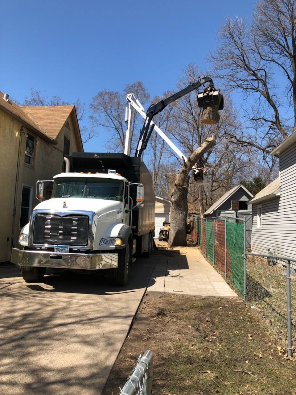 A tree removal service truck with a crane cutting and loading tree branches in a narrow driveway.