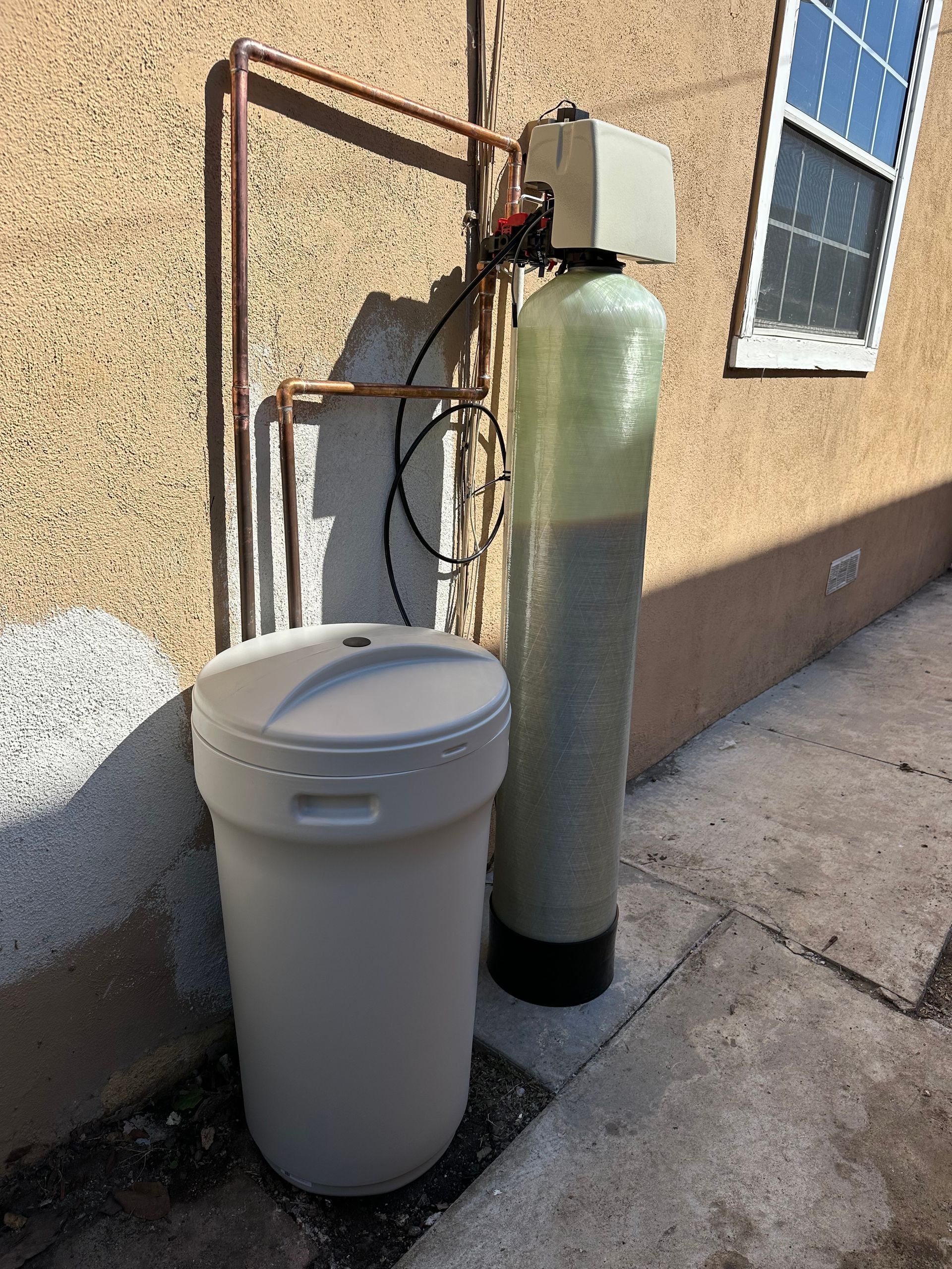 Water softener system next to a salt storage tank, near a building wall and window.