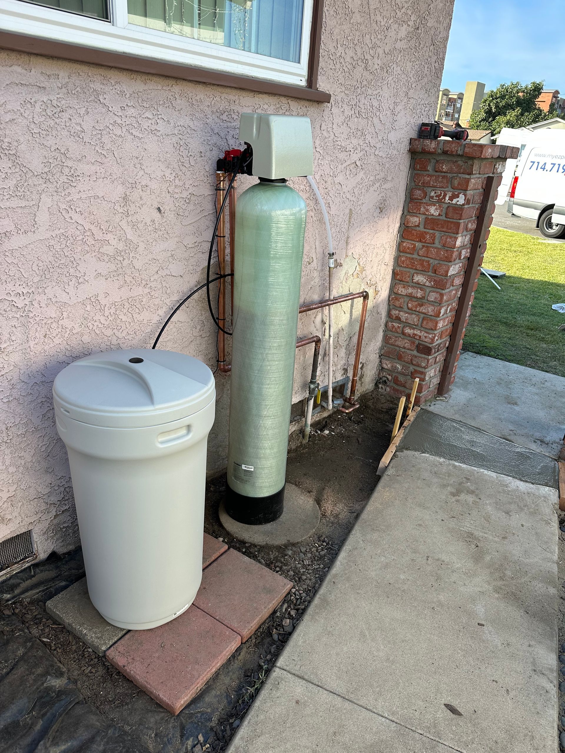 Water softener system outside a house with a pink stucco wall and a brick pillar.