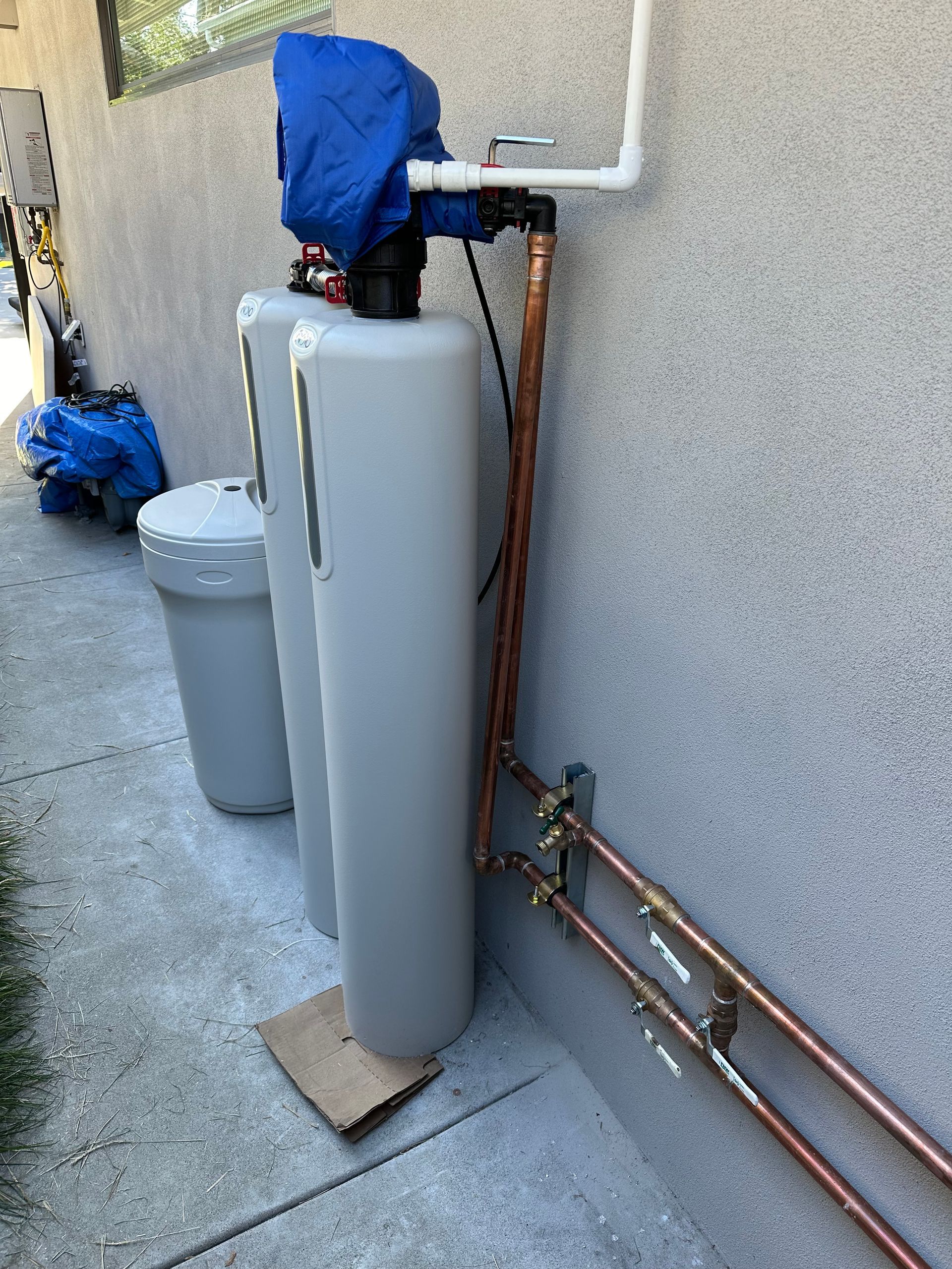 Water softener system with tanks, copper pipes, and a blue cover outside on a concrete wall.