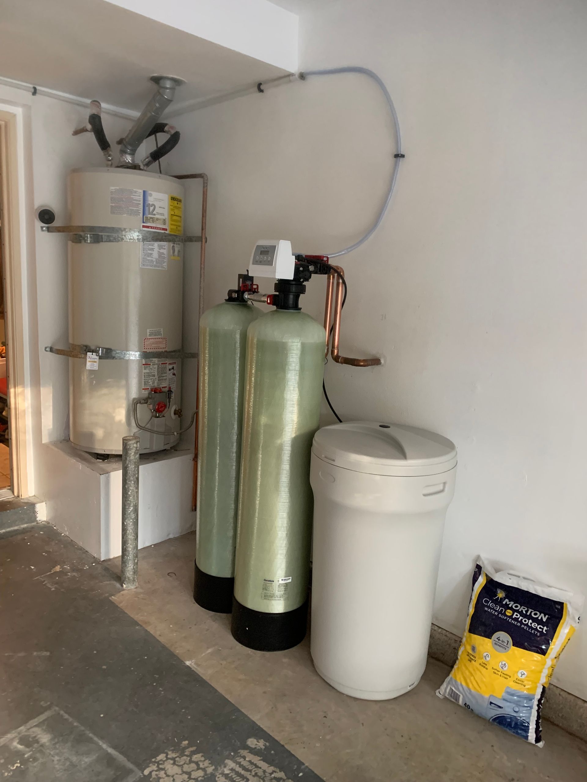 Water softener system and water heater in a garage; tanks, salt bag, and pipes visible.