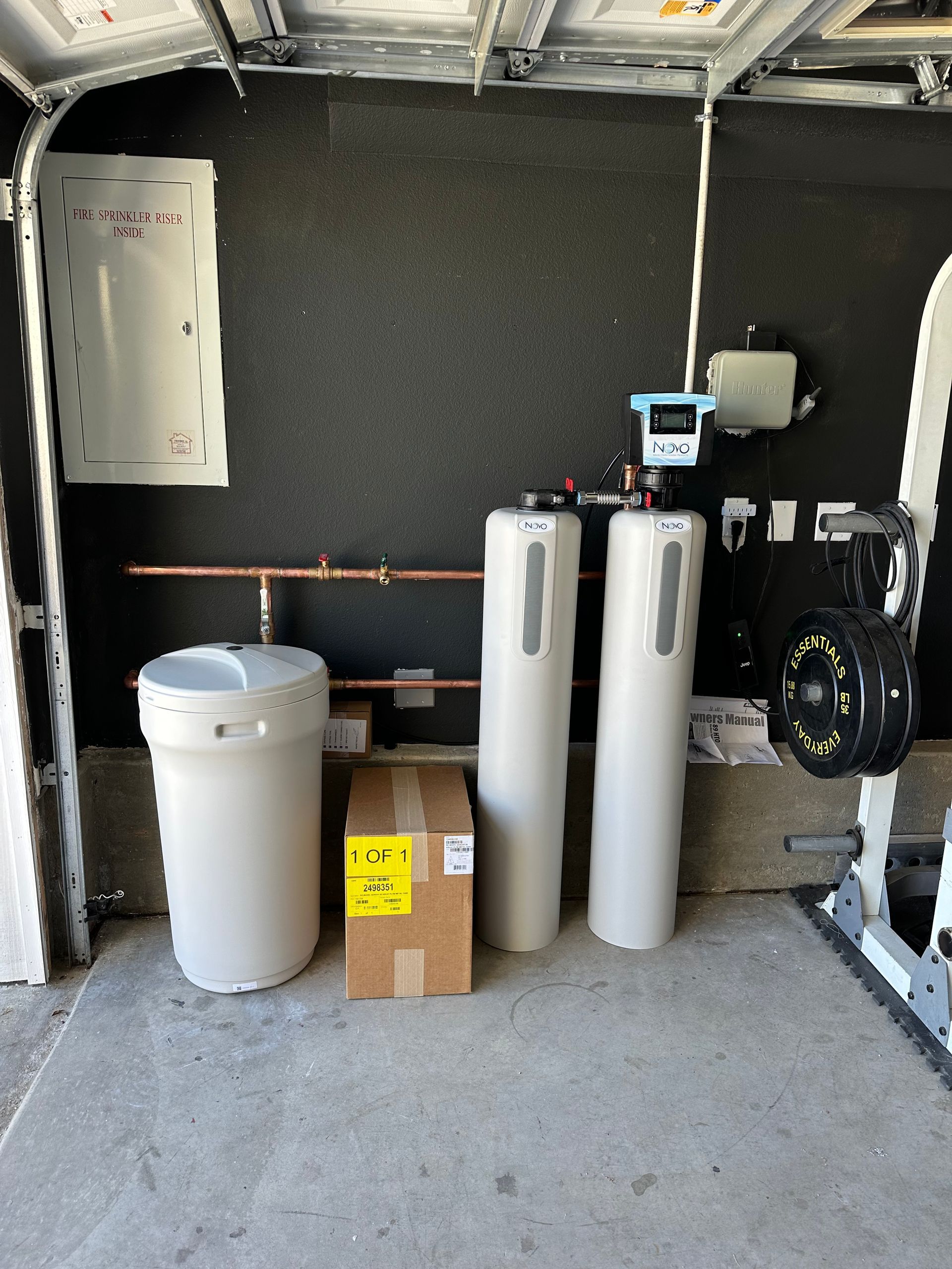 Water softener system in a garage: tank, two cylinders, and control panel against a dark wall.