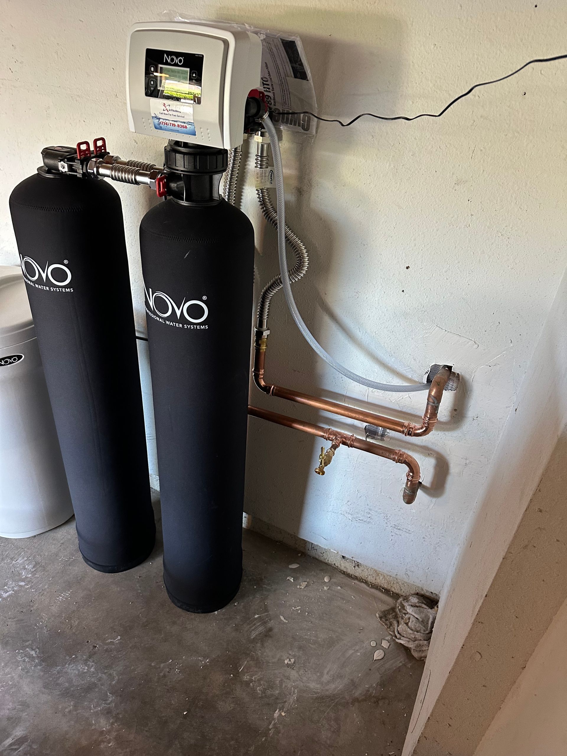 Water filtration system, black tanks and white control panel, installed on a concrete wall in a utility room.
