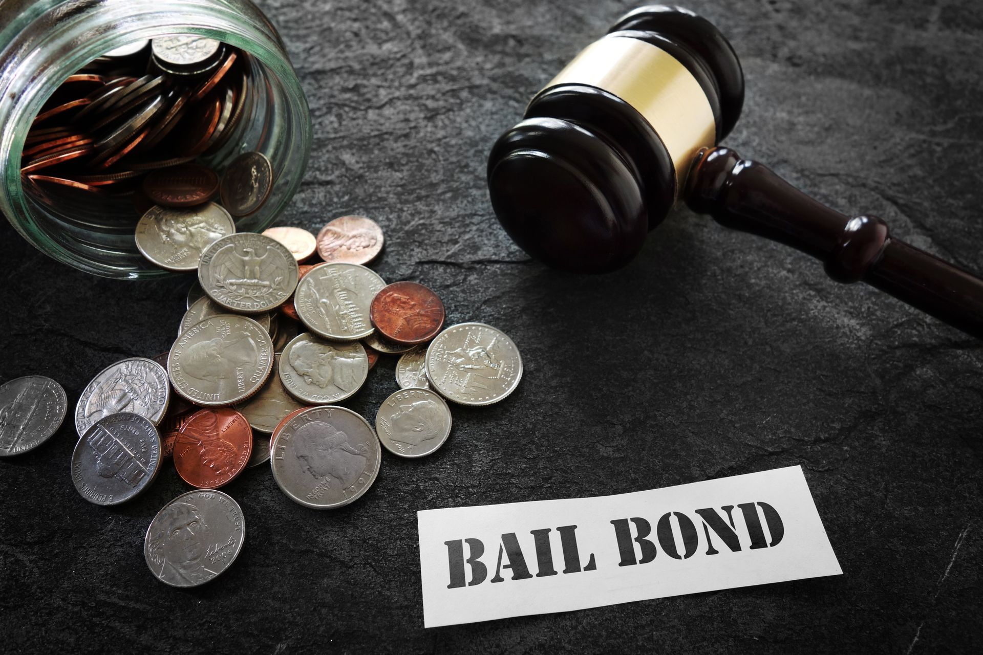 A judge's gavel rests next to a spilled jar of coins and a sign that reads BAIL BOND on a dark, textured surface.