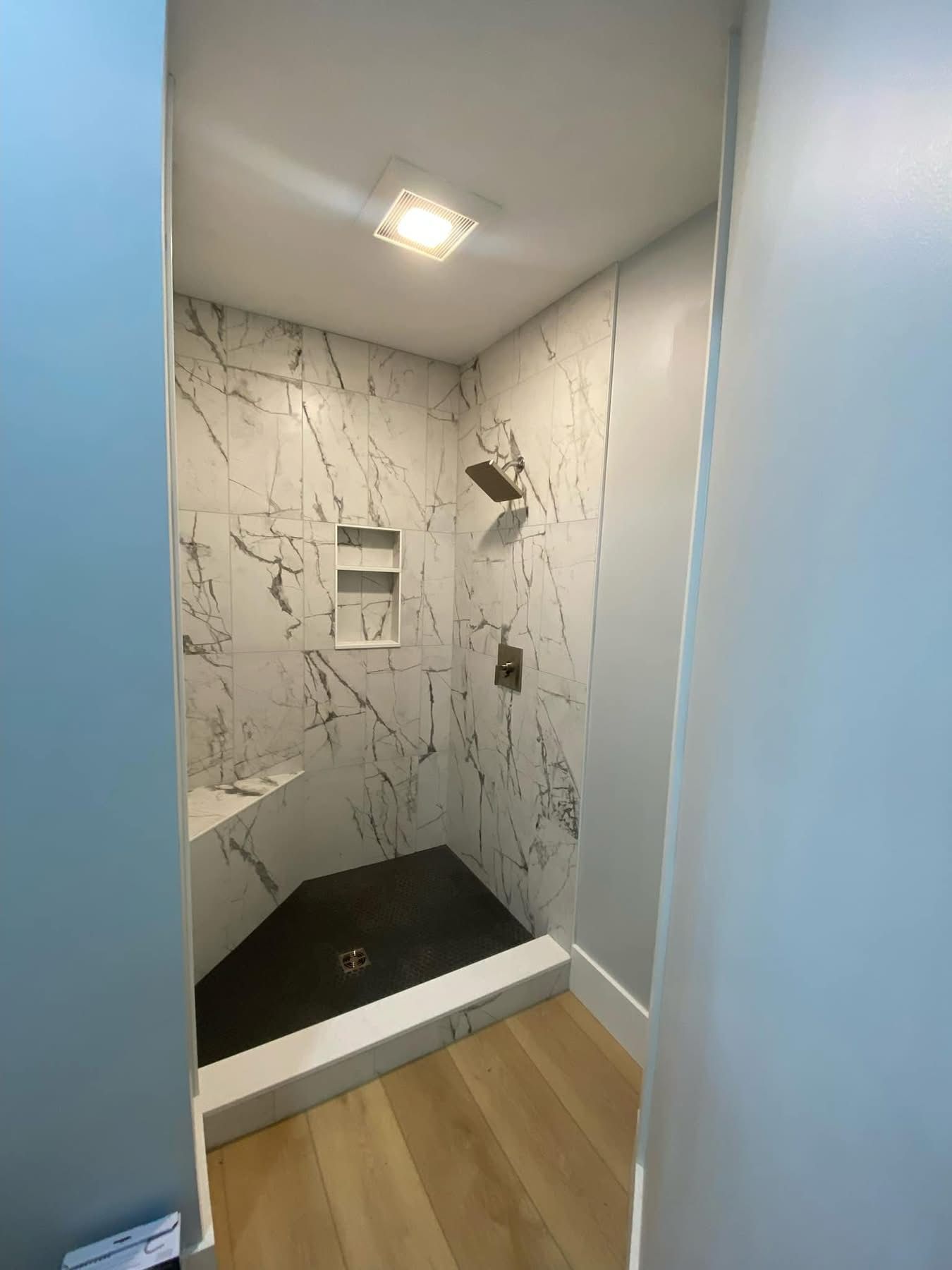 A walk-in shower with white marble-patterned tile walls, a dark stone floor, a built-in bench, and a recessed wall niche.