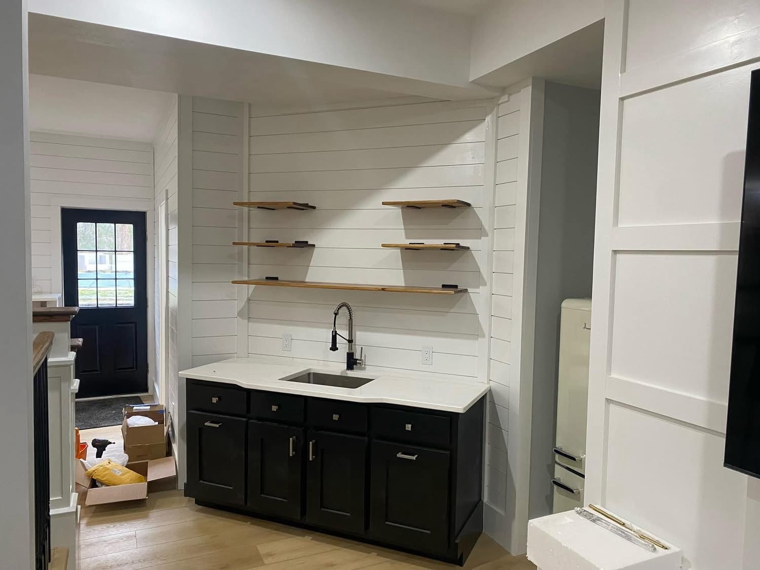 A wet bar with black cabinets, white shiplap walls, three floating wood shelves, and a faucet against a white countertop.