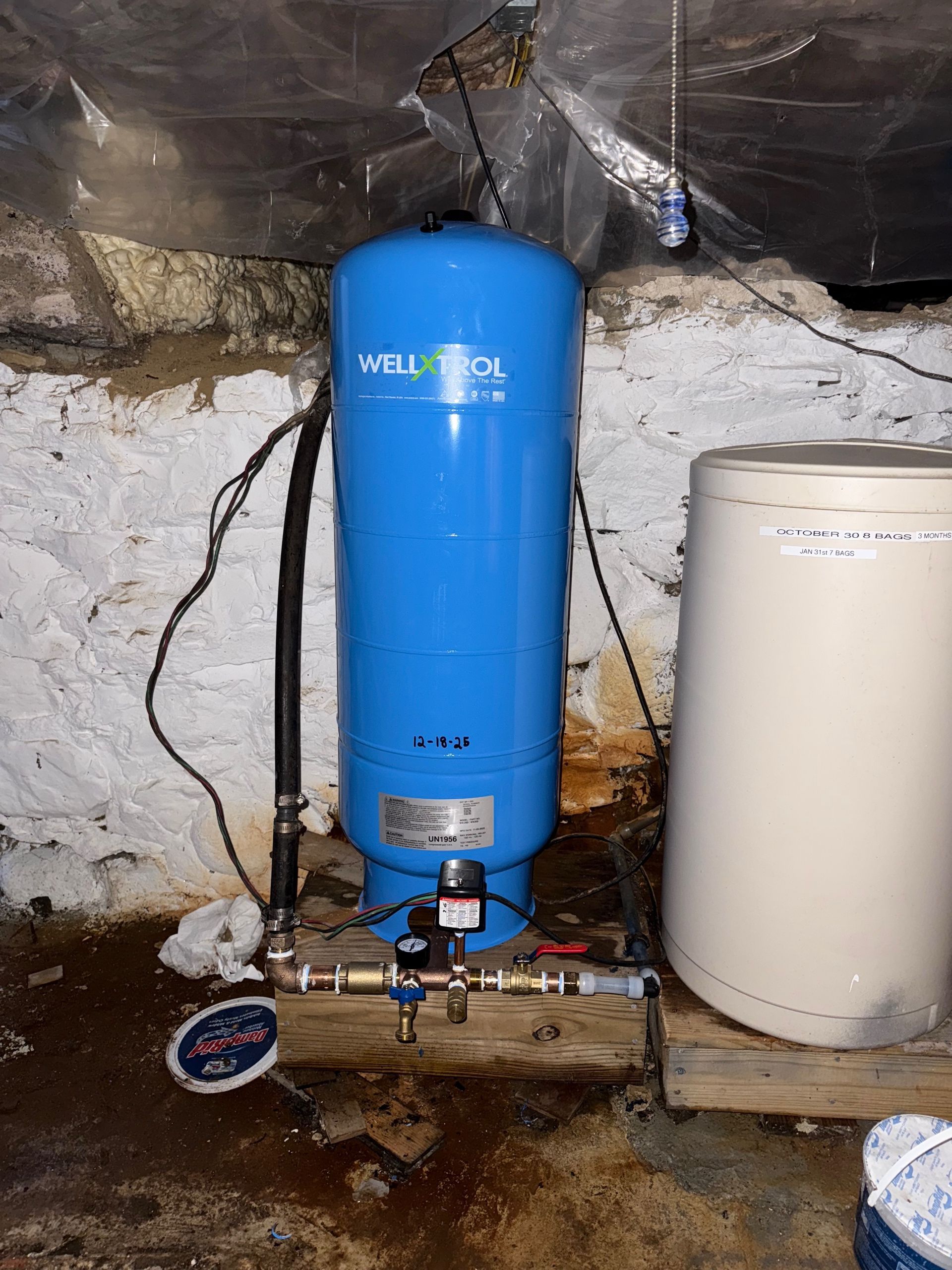A blue Well-Trol water pressure tank and a beige water treatment tank stand on a wooden platform in a basement.