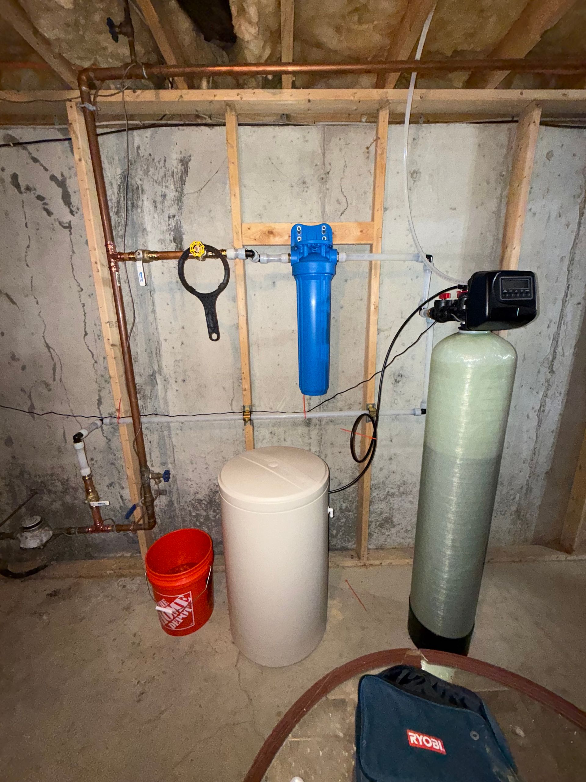 A home water filtration and softening system with two tall tanks, a beige salt brine tank, and a blue filter against a wall.