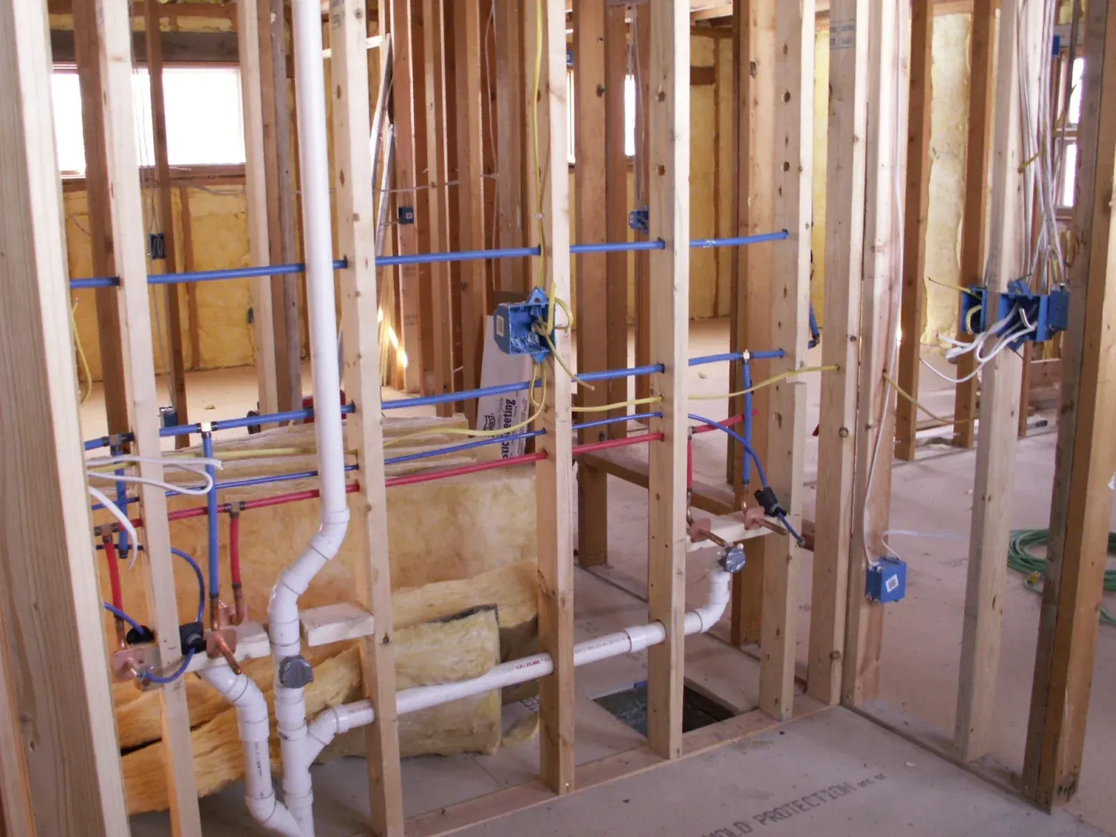 Construction framing of an interior wall showing installed white PVC plumbing and blue and red PEX water lines.