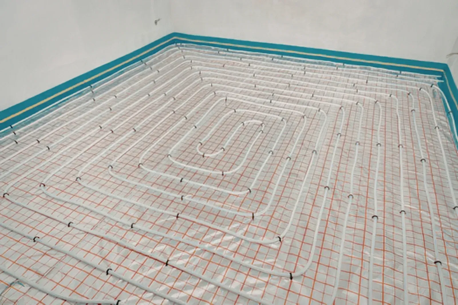 White radiant floor heating pipes installed in a spiral pattern over a grid-patterned underlayment in a room.