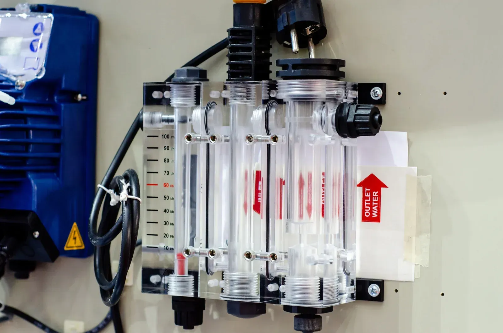 A wall-mounted industrial flow meter system with three transparent tubes and a blue pump component on the left.