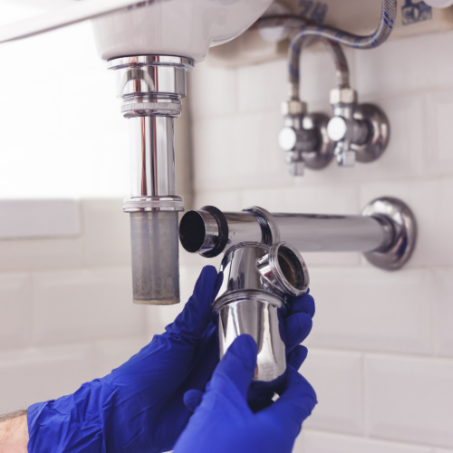 A person wearing blue nitrile gloves holds the removed P-trap of a chrome sink drain pipe assembly.