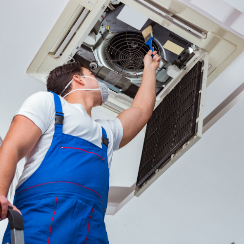A worker in blue overalls and a face mask cleaning a ceiling-mounted air conditioning unit with a brush.