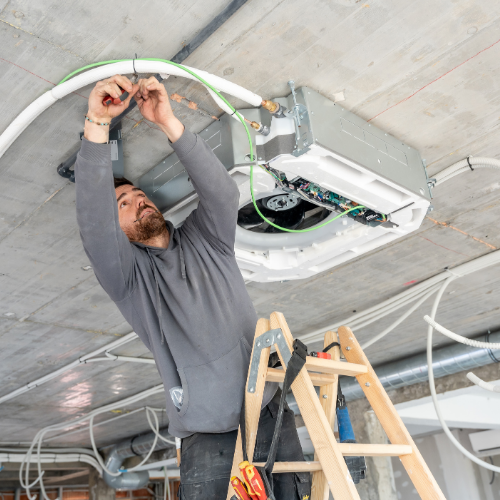 A person on a stepladder installs wiring inside an HVAC unit mounted to an unfinished concrete ceiling.