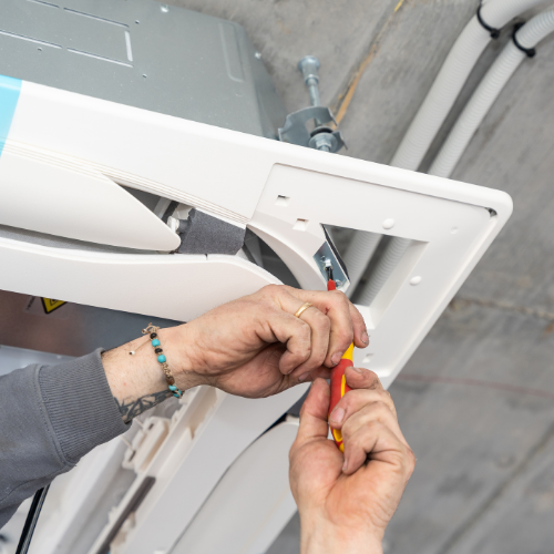 Close-up of hands using a screwdriver to tighten a screw on a white HVAC unit mounted to a concrete ceiling.
