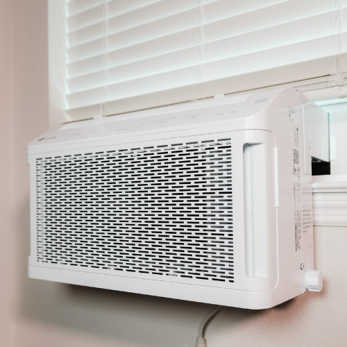 A white window-mounted air conditioner installed in a room with white blinds.