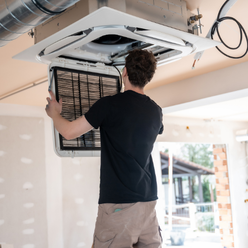 A technician in a black shirt and tan pants services a ceiling-mounted HVAC unit by removing its filter.