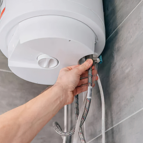 A hand adjusts a metal braided water hose connected to the bottom of a white electric water heater mounted on a tiled wall.