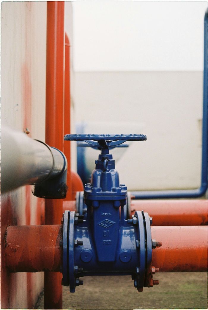 A blue gate valve installed on an orange industrial pipe against a white wall.