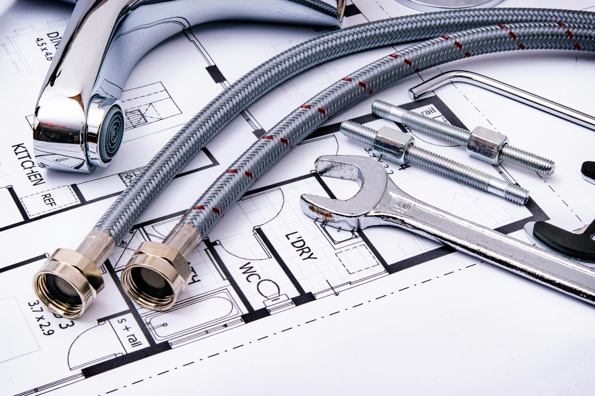 A metal faucet, braided water supply lines, and a wrench arranged on top of a house floor plan blueprint.
