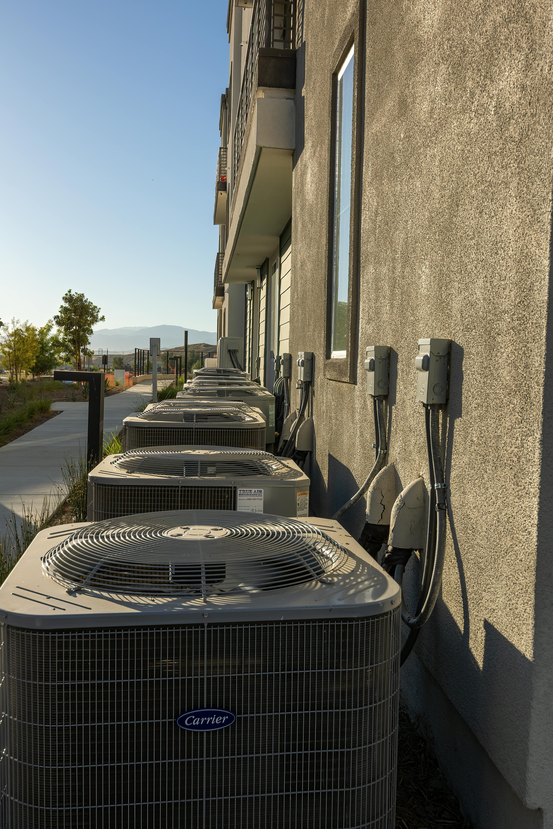 A row of outdoor air conditioning units sits along the exterior stucco wall of a multi-story residential building.