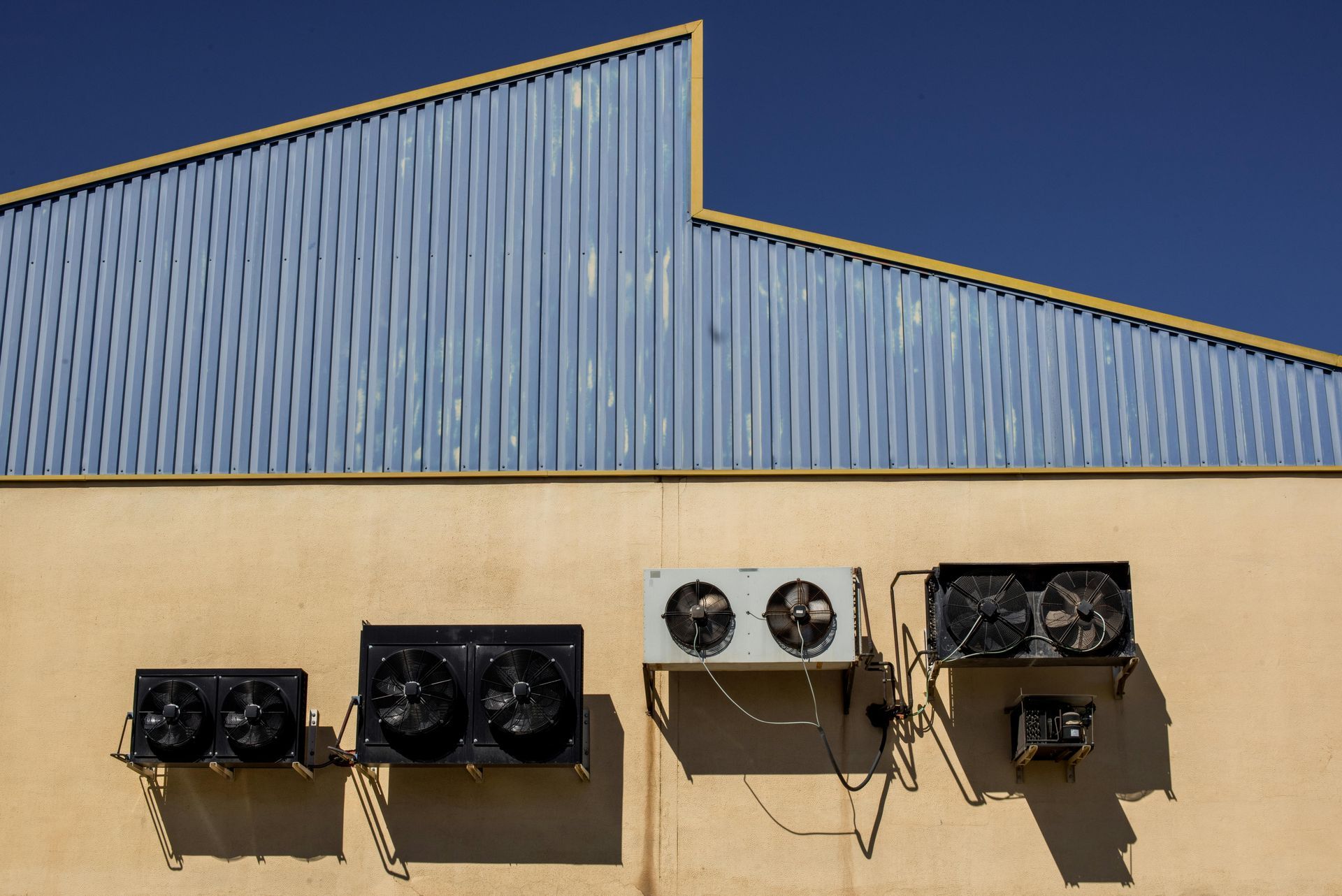 Four industrial cooling units mounted on a tan concrete wall beneath a blue corrugated metal roof against a clear sky.