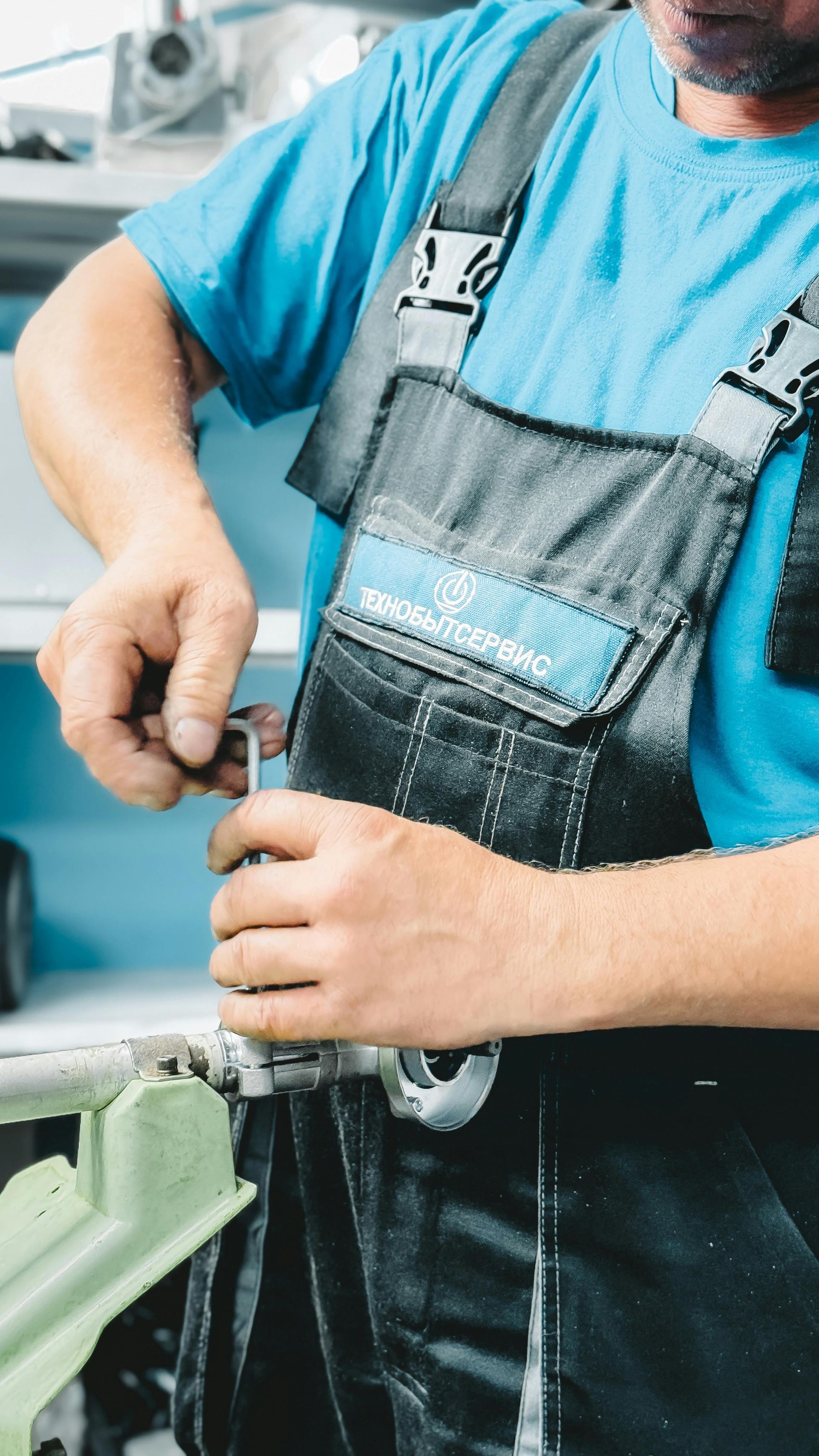 A worker in blue shirt and dark overalls uses an L-shaped hex key to tighten a bolt on a metal component in a workshop.