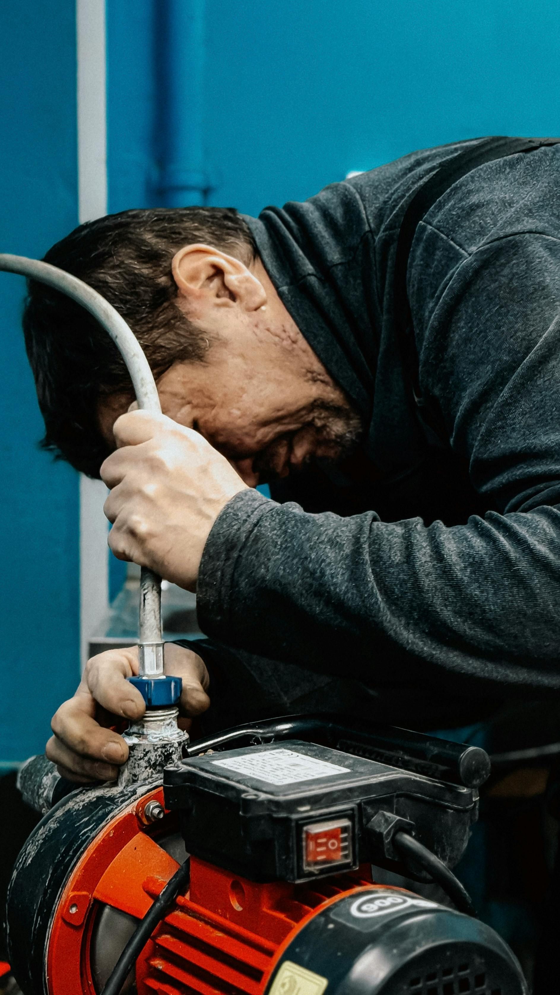 A person focused on connecting a gray hose to the top of a red electric pump against a bright blue wall.
