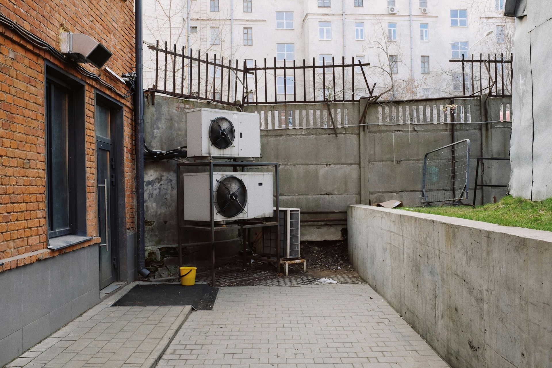 Two stacked air conditioning units sit on a metal frame against a concrete wall near a brick building.